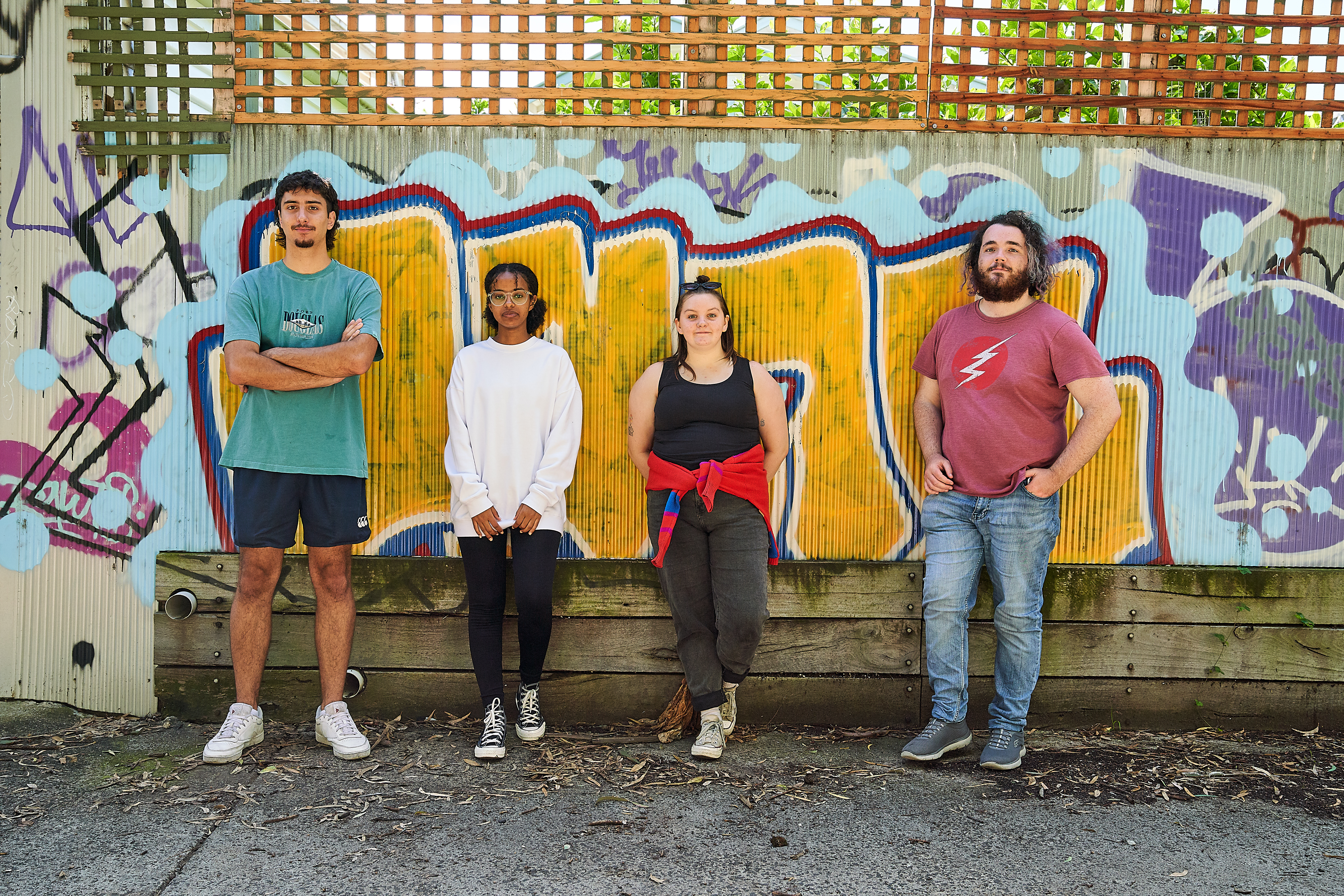 Four young people standing in front of a graffitied wall looking at the camera